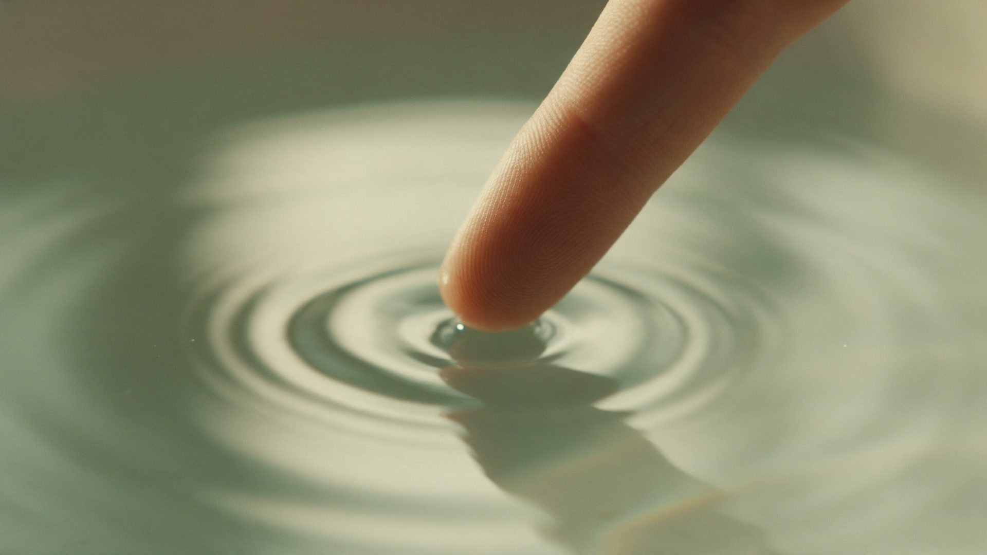 A fingertip touching still water, sending out rings of ripples.